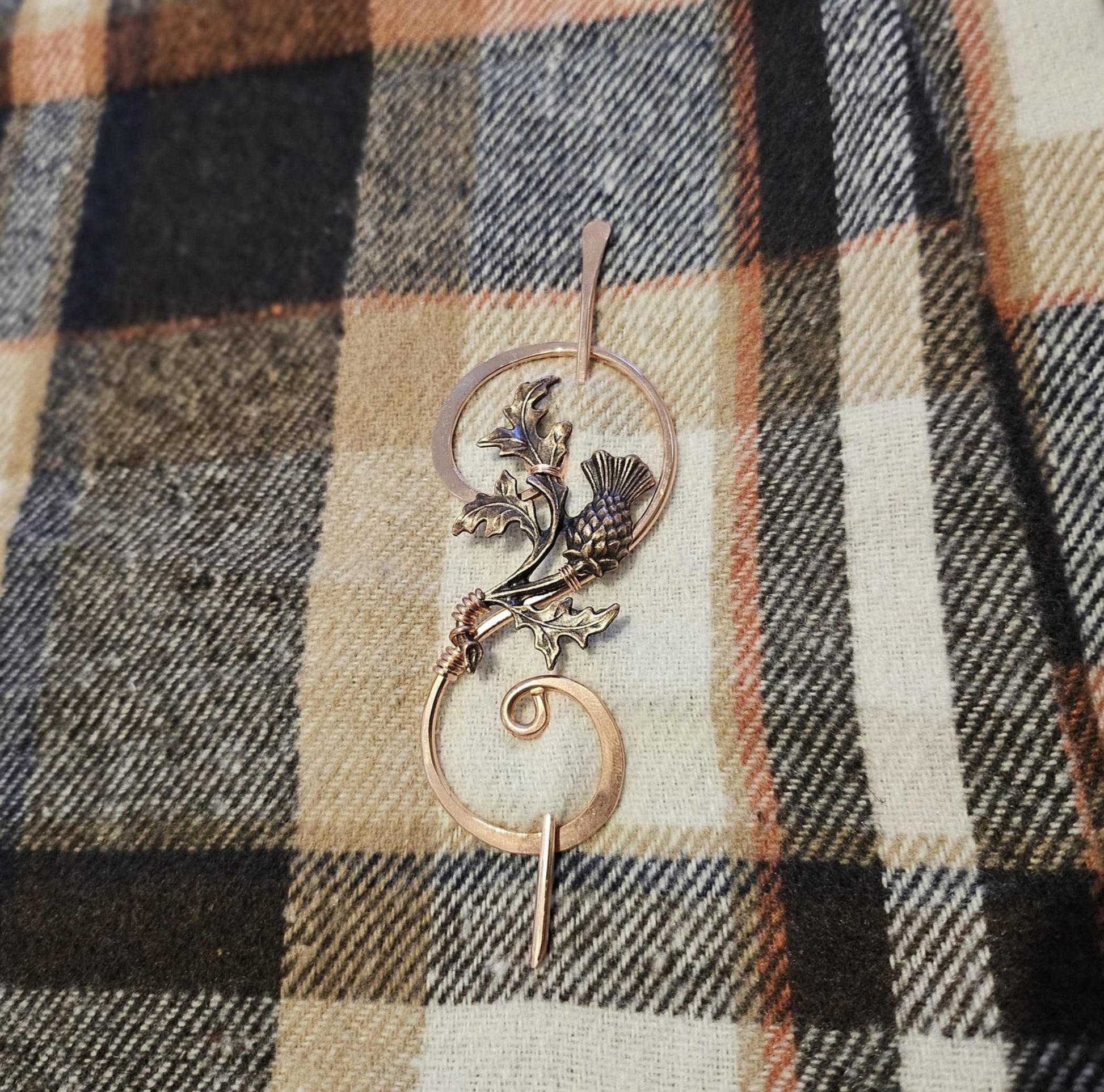 Solid copper thistle shawl pin shown pinned through a traditional wool tartan scarf, featuring handcrafted wire-work and a tapered focal pin.