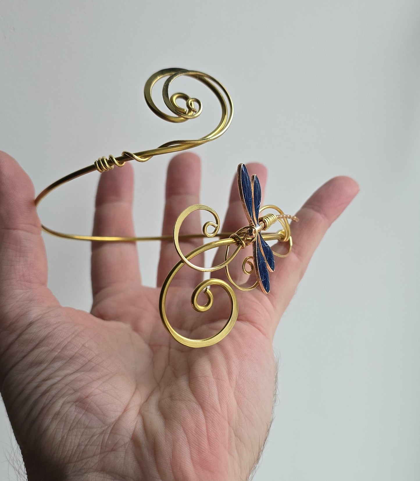 Handheld view of a bespoke gold-finish brass armlet, showing the dragonfly's iridescent-style blue wings and hand-formed wire curls. This scale-reference photo highlights the sturdy, adjustable design and the meticulous craftsmanship of the elven-inspired jewelry.