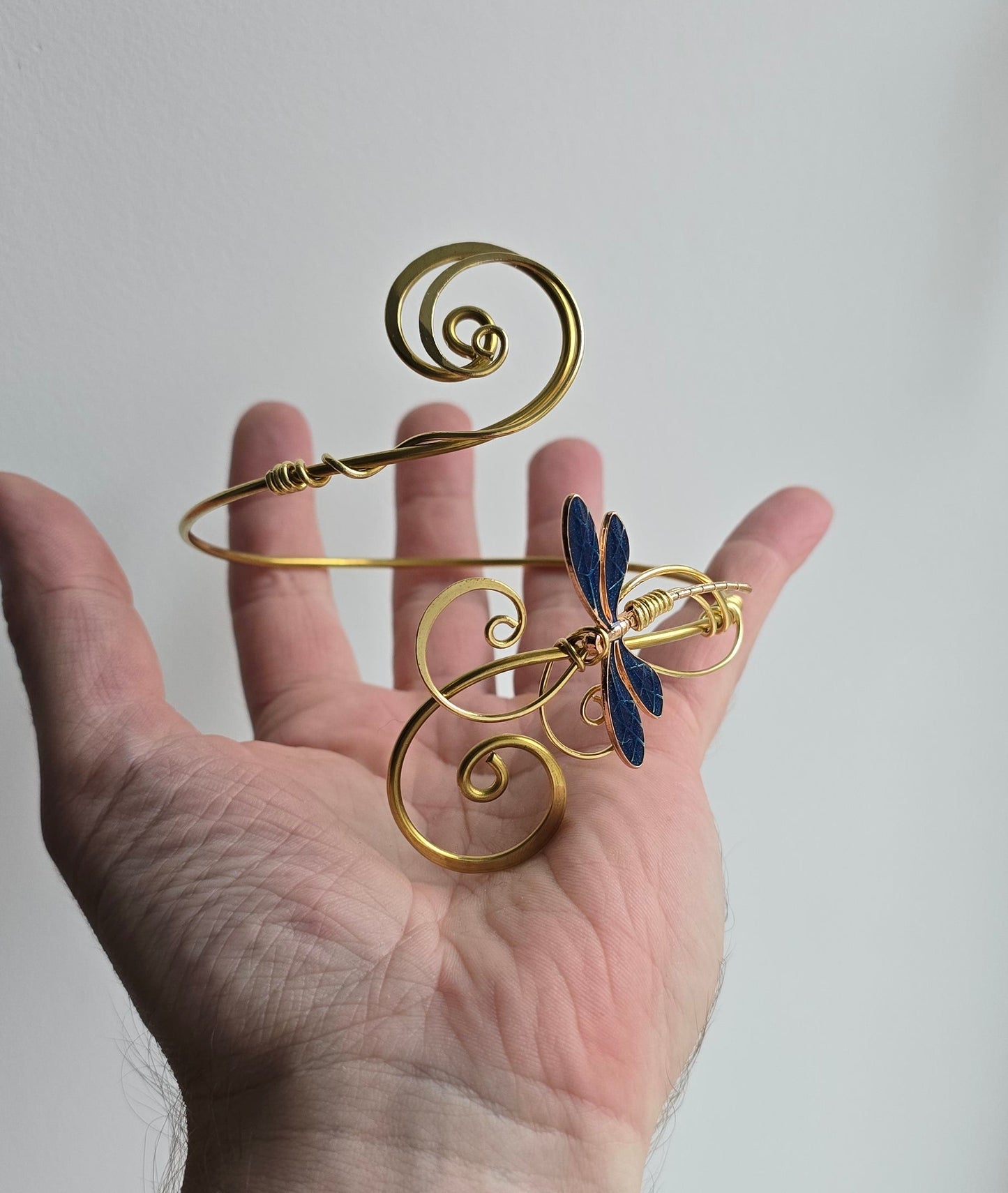 A close-up photo showing a hand holding the gold brass dragonfly arm cuff to demonstrate scale. The image highlights the sturdy wire-wrapped construction and the delicate blue dragonfly wings against a neutral light background.