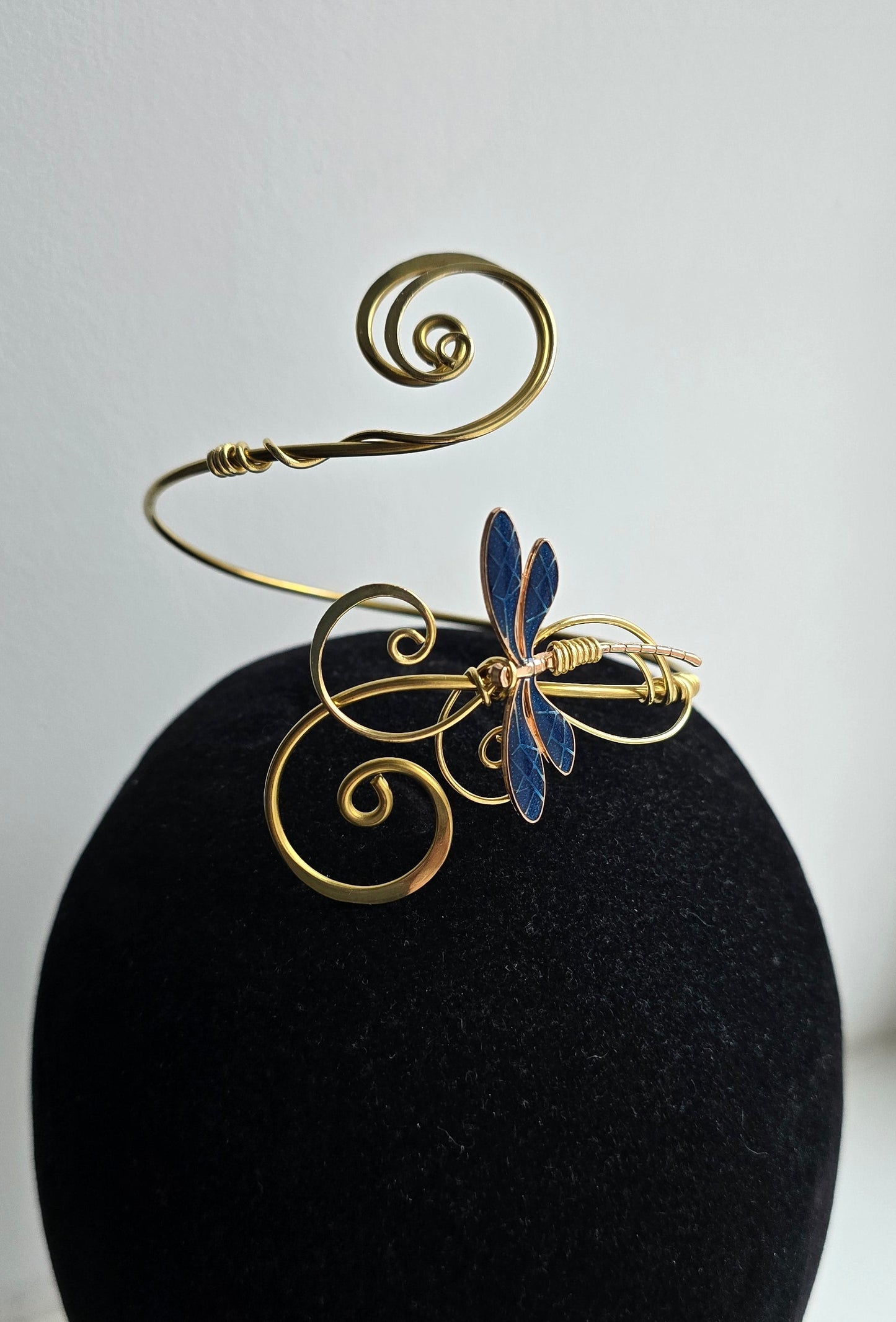 Detailed close-up of a handcrafted brass wire armlet featuring a central blue dragonfly motif with intricate spiral filigree, presented on a black jewelry form to highlight the metallic gold luster and textured enamel wings.
