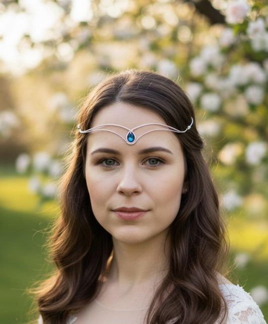 A model wearing a handcrafted silver elven forehead crown with a Bermuda Blue crystal droplet. Styled for a fantasy wedding or ethereal cosplay in a sunlit garden setting, showing the delicate woven wire design and forehead fit