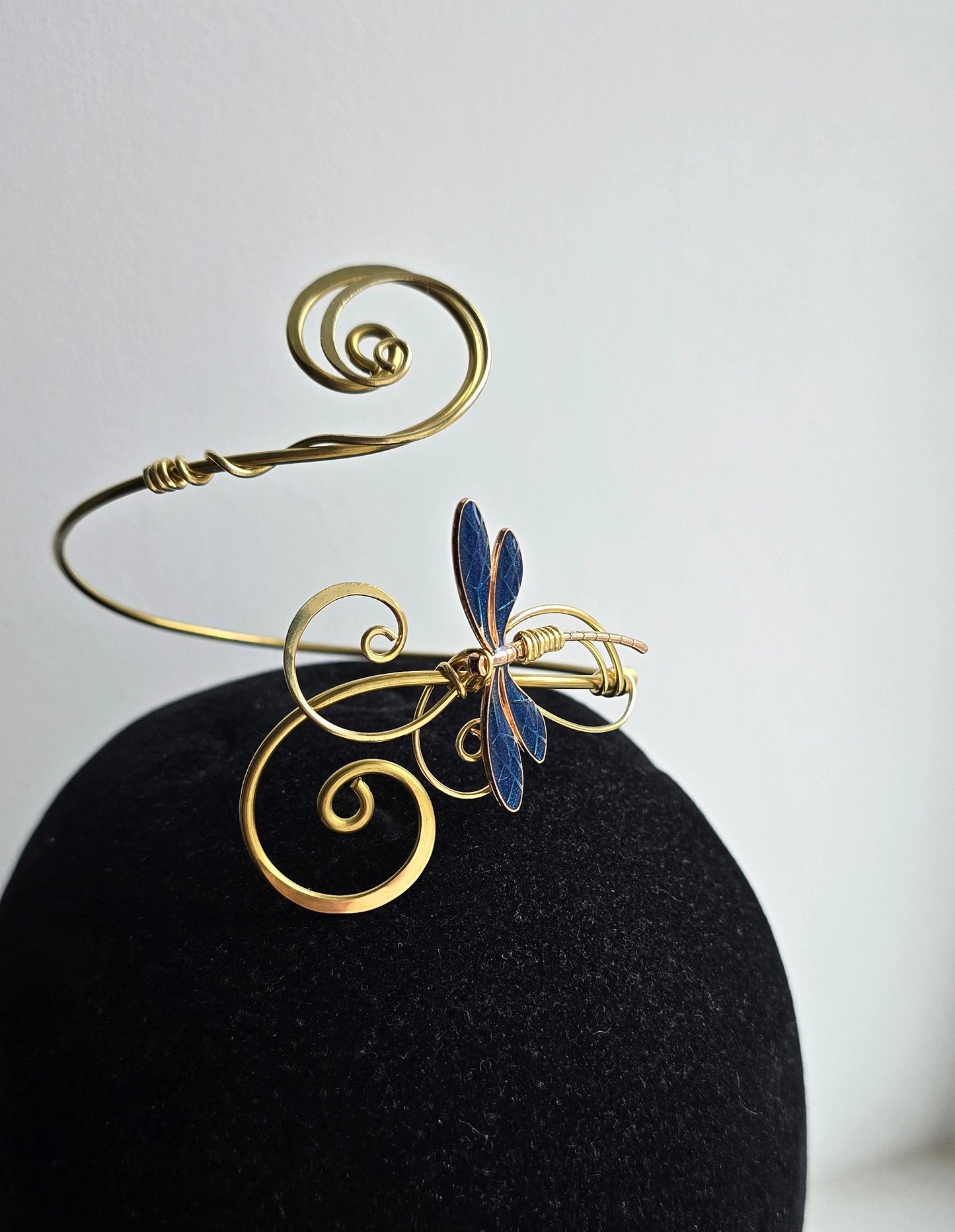 A sculptural gold brass dragonfly arm cuff with curled filigree accents displayed on a black velvet stand. The intricate blue dragonfly wings stand out against the high-contrast dark background, showcasing the handcrafted wire-work.
