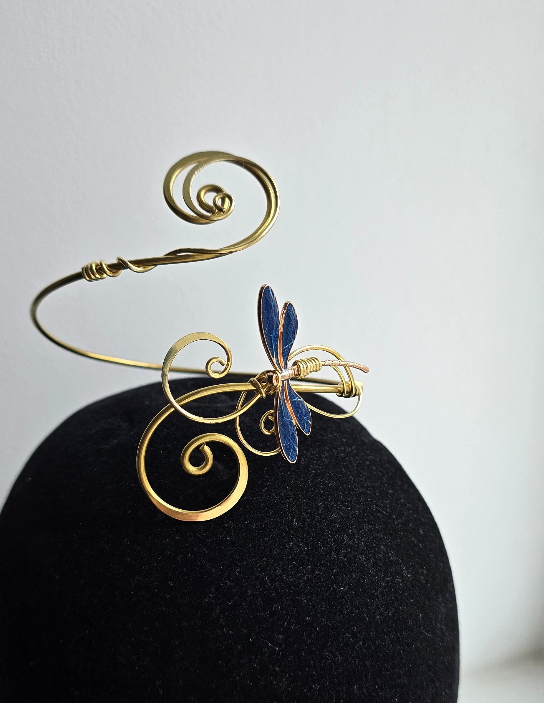 A sculptural gold brass dragonfly arm cuff with curled filigree accents displayed on a black velvet stand. The intricate blue dragonfly wings stand out against the high-contrast dark background, showcasing the handcrafted wire-work.