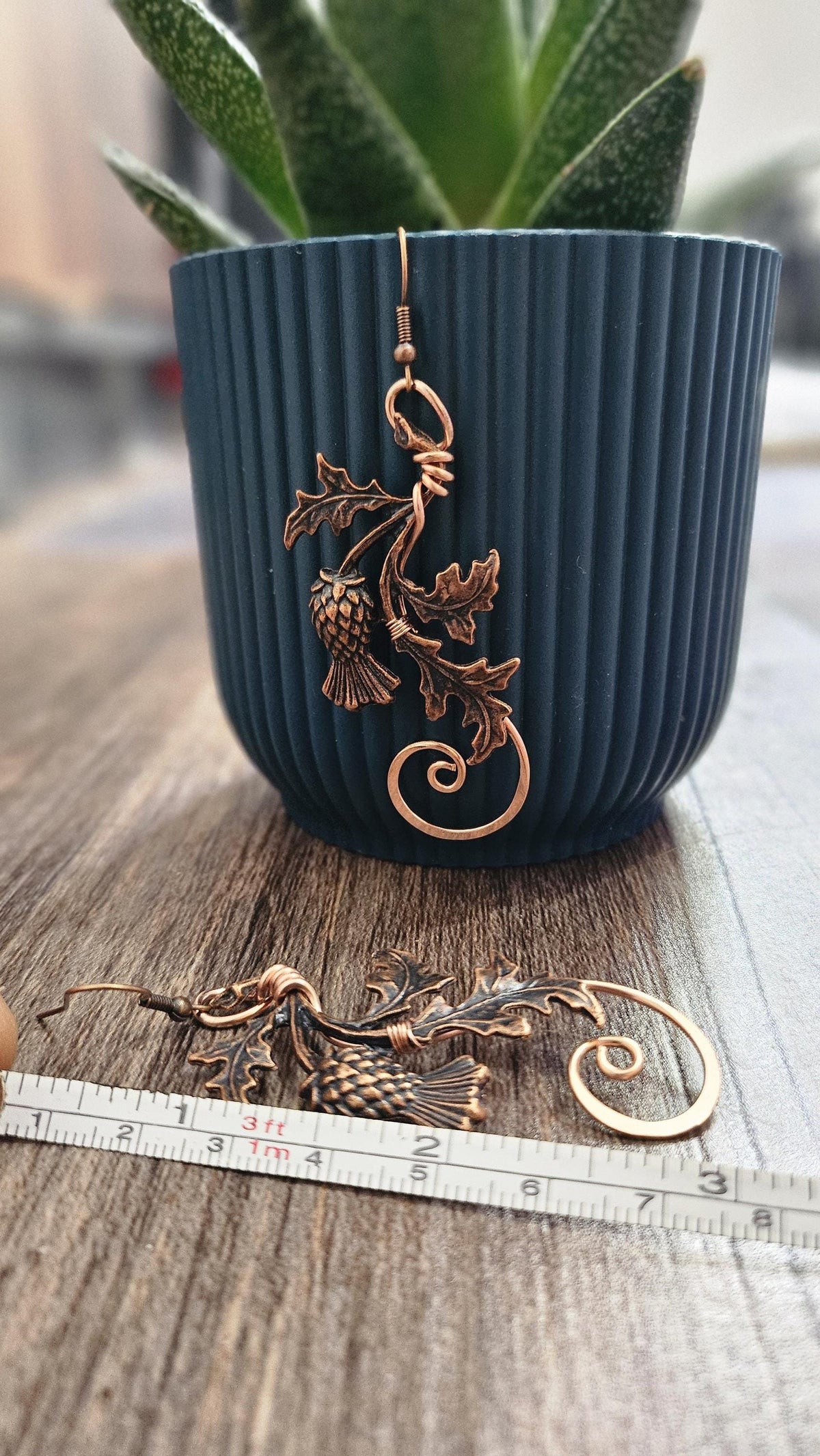 Handmade copper wire Scottish Thistle earrings hanging from a blue plant pot, showing the intricate metalwork and 3-inch drop length.