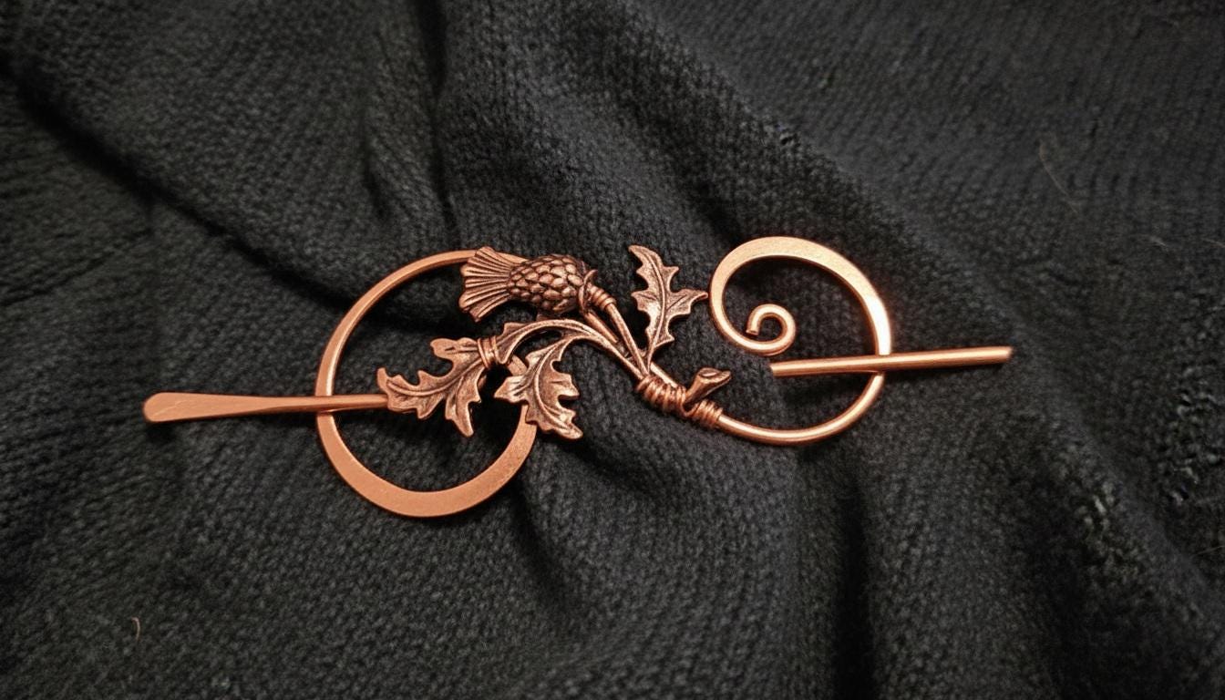 Solid copper thistle shawl pin shown pinned through a traditional wool scarf, featuring handcrafted wire-work and a tapered focal pin.