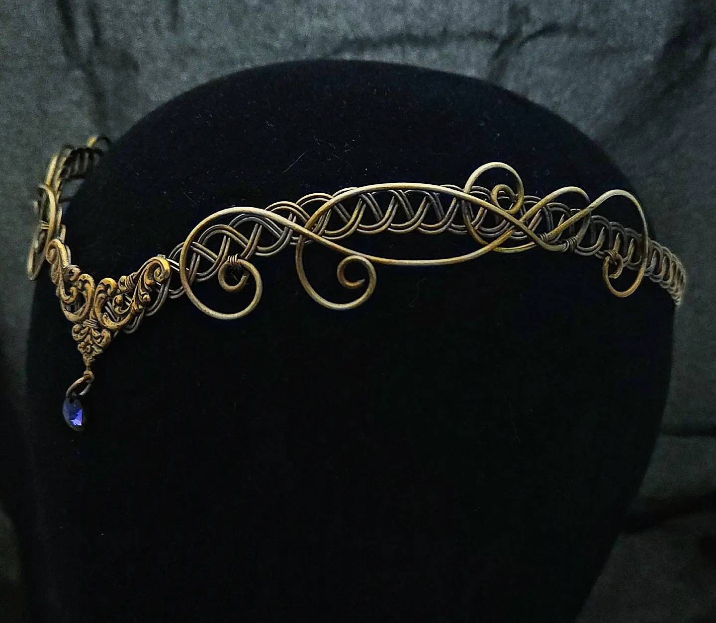 Detailed side view of a handcrafted antique brass medieval circlet featuring a Celtic weave and a purple heliotropic crystal drop on a black display.