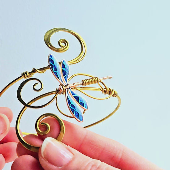 Macro video of a gold-finish brass dragonfly arm cuff being held to show the shimmering blue wing details and hand-curled wire filigree. The video highlights the high-shine metallic finish and the intricate handcrafted texture of the elven-style jewelry.