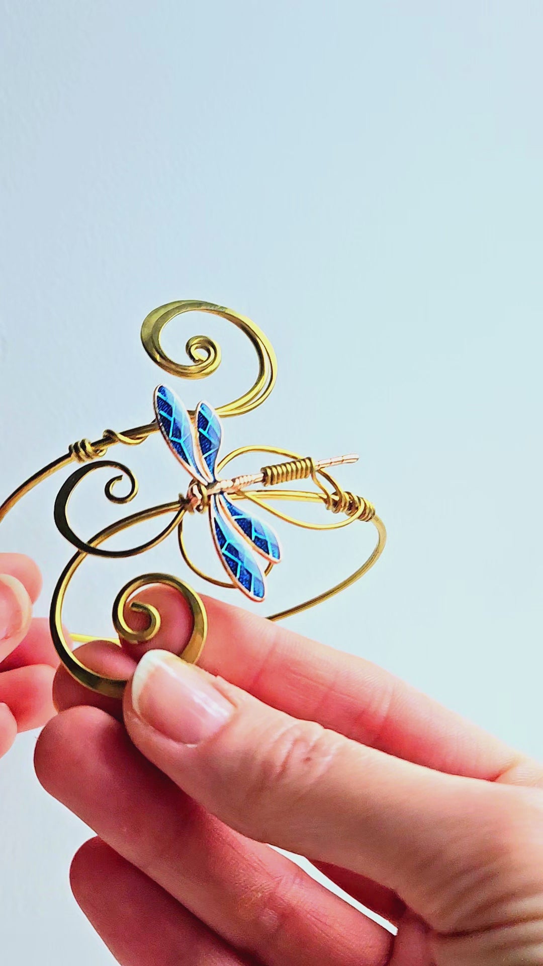 Macro video of a gold-finish brass dragonfly arm cuff being held to show the shimmering blue wing details and hand-curled wire filigree. The video highlights the high-shine metallic finish and the intricate handcrafted texture of the elven-style jewelry.