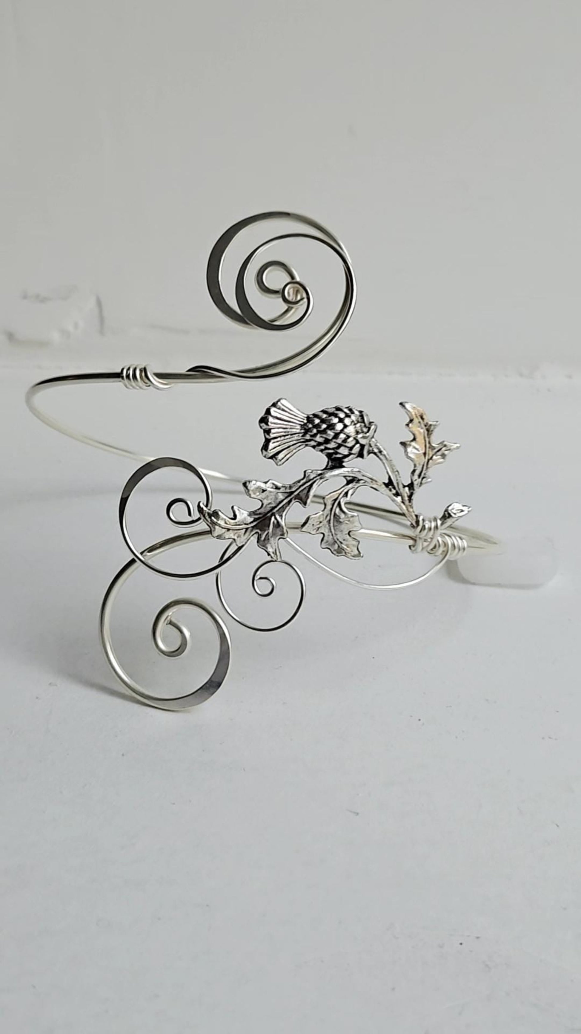 Handcrafted silver-plated thistle arm cuff featuring an intricate wire-wrapped scroll design and a detailed thistle charm on a light background.